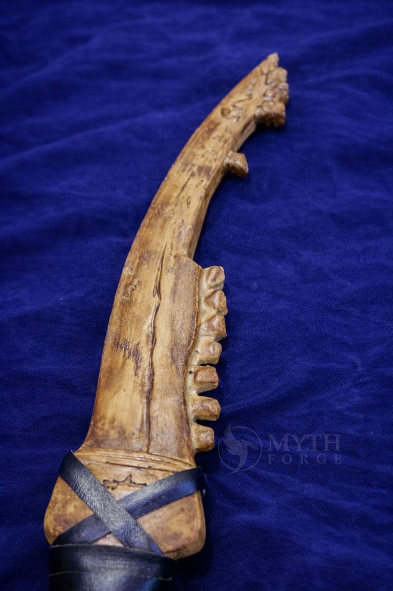 May include: A carved, bone-colored dagger with a curved blade and a textured handle. The handle is wrapped with black leather straps. The dagger has a unique, rustic appearance, suggesting a handmade or ancient origin. The Myth Forge logo is visible.