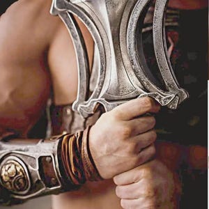 May include: A close-up of a silver sword with an ornate handle, held by a person. The sword has a detailed design. The person is wearing a brown leather arm guard with gold accents.