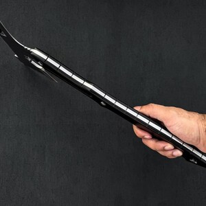 May include: A black and silver tool with a curved blade and a long, segmented handle. The tool is held in a hand against a dark background. The blade has a triangular tip.