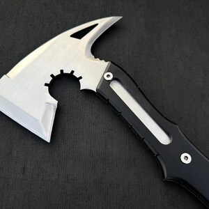 May include: A silver and black tactical axe with a sharp blade and a curved top. The axe features a serrated edge and a black handle with a white accent. The design is modern and functional, suitable for outdoor or survival use.