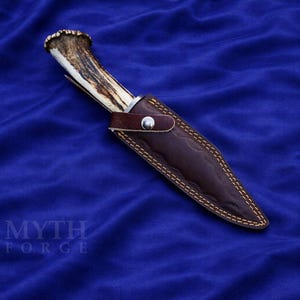 May include: A hunting knife with an antler handle and a brown leather sheath. The sheath is stitched with yellow thread and secured with a silver button. The knife is set against a blue background.
