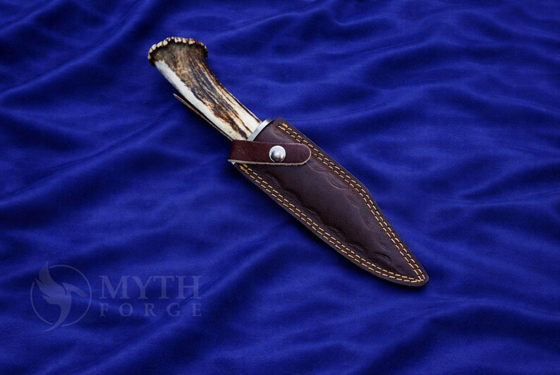 May include: A hunting knife with an antler handle and a brown leather sheath. The sheath is stitched with yellow thread and secured with a silver button. The knife is set against a blue background.