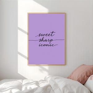 Peut inclure: Une affiche encadrée avec un fond violet clair et les mots "sweet sharp iconic" en écriture noire. L'affiche est dans un cadre en bois clair et est affichée sur un mur blanc au-dessus d'un lit avec des oreillers.
