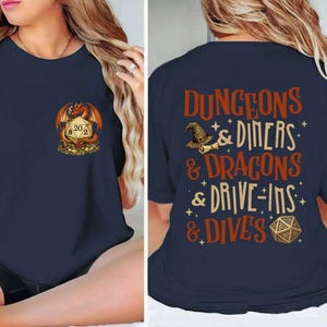 May include: Navy blue t-shirt featuring a dragon and dice graphic on the front, and text on the back that reads "DUNGEONS & DINERS & DRAGONS & DRIVE-INS & DIVES". The shirt has a round neckline and short sleeves.