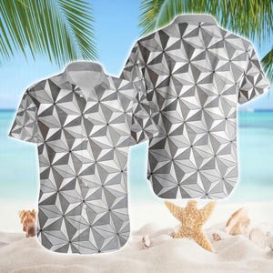 May include: A short-sleeved button-down shirt with a geometric pattern in shades of gray and white. The shirt is displayed on a beach setting, with a second shirt visible in the background. The design features a repeating pattern of three-dimensional shapes.