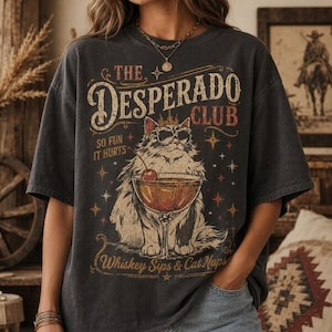 May include: Dark gray t-shirt with a graphic of a cat wearing sunglasses and a crown, holding a cocktail. Text reads "The Desperado Club" and "Whiskey Sips & Cat Naps". The shirt has a vintage, distressed look.