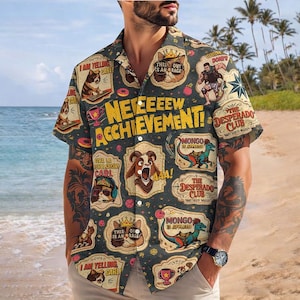 May include: Short-sleeved button-down shirt with a dark gray background and various cartoon graphics. The shirt features text that reads "NEEEEEW ACHIEVEMENT!" and other phrases. The design includes images of cats, dinosaurs, and other characters.