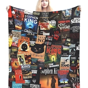 May include: A black blanket featuring a collage of Stephen King book covers. The covers display various colors and designs, with titles like "The Shining" and "Dead Zone". The blanket is held up by a person.