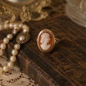 May include: An oval-shaped gold ring featuring a carved shell cameo of a profile portrait. The cameo displays a white figure against a burnt orange background, set within a polished gold metal frame with a simple band design.