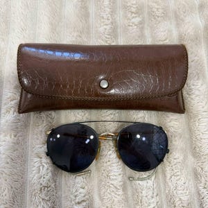 May include: Sunglasses with dark lenses and a gold-colored frame, paired with a brown leather case. The case has a button closure and a textured surface. The sunglasses have a classic, round shape.