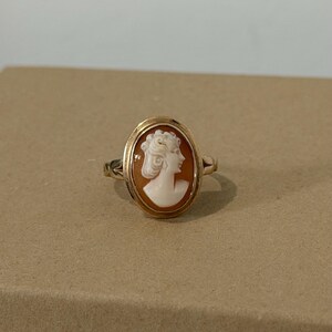 May include: A gold ring featuring an oval cameo. The cameo depicts a woman's profile in white against a reddish-brown background. The ring has a delicate, vintage design.