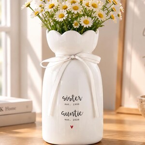 May include: A white ceramic vase with a scalloped rim and a light beige ribbon tied around the neck. The vase features black text reading "sister est. 1999" and "auntie est. 2026" with a small red heart below. It is filled with a bouquet of white daisies.