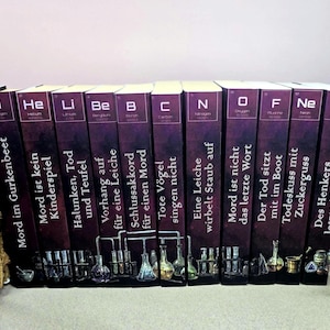 Könnte beinhalten: Eine Sammlung von Hardcover-Büchern mit violetten Buchrücken, die jeweils mit einem chemischen Elementsymbol und einem deutschen Titel versehen sind. Die Bücher zeigen Illustrationen von Laborgeräten am unteren Rand. Titel sind u.a. "Mord im Gurkenbeet" und "Todeskuss mit Zuckerguss".