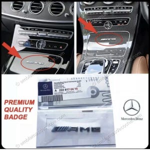 Puede incluir: La imagen muestra un emblema AMG plateado, instalado en el interior de un coche y como producto separado. El emblema se muestra sobre una superficie negra brillante y un panel de fibra de carbono. También se ve el logotipo de Mercedes-Benz y el texto "PREMIUM QUALITY BADGE".