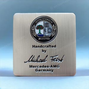 May include: A square, brushed metal plaque featuring the Mercedes-AMG logo at the top. The logo is a circular emblem with a tree, a gear, and the text "AMG". Below the logo, the text reads "Handcrafted by Michael Feest, Mercedes-AMG Germany."