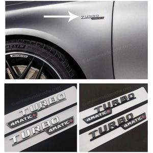 Puede incluir: Emblema de coche "TURBO 4MATIC" plateado y negro. El emblema se muestra en un coche gris y en una tarjeta blanca. La rueda del coche es visible en el fondo. El emblema tiene un acento rojo.