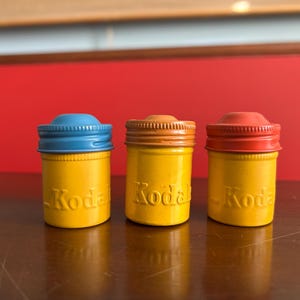 Może przedstawiać: Trzy zabytkowe pojemniki na film Kodak. Każdy pojemnik jest ż&oacute;łty z kolorową pokrywką: niebieską, pomarańczową i czerwoną. Słowo "Kodak" jest wytłoczone na przodzie każdego pojemnika. Tło to czerwona ściana.