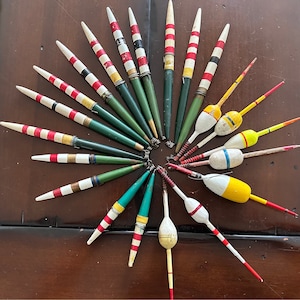 May include: A collection of 20 vintage wooden fishing bobbers in various colors, including red, white, green, yellow, and black. The bobbers are arranged in a circular pattern on a wooden surface.