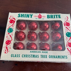 May include: A box of twelve Shiny Brite glass Christmas tree ornaments in a red metallic finish. The rectangular cardboard packaging features green and red text reading Shiny Brite and American Made Glass Christmas Tree Ornaments.