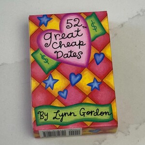 May include: A rectangular box with the title "52 Great Cheap Dates" on the front. The box has a colorful design with a pink heart, blue stars, and green dollar signs. The author's name, "By Lynn Gordon," is printed at the bottom.