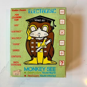 May include: Vintage Radio Shack "Monkey See" electronic learning game. The box features a cartoon monkey wearing a graduation cap, with math symbols and numbers. The text includes "Add", "Subtract", "Multiply", "Divide", and "Games". The box is green, brown, and yellow.