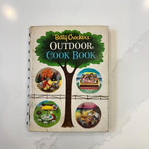 May include: Vintage Betty Crocker's Outdoor Cook Book with a spiral binding. The cover features a tree design with the title in green and white. Four circular illustrations depict outdoor cooking scenes, including a picnic and a campfire.