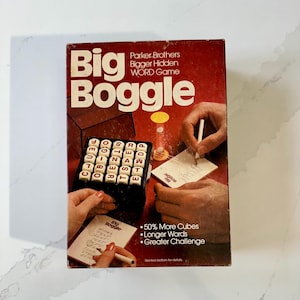 May include: A vintage Big Boggle word game box. The red box features the game title in large white letters. The box shows the game in play, with hands writing on notepads and the game board visible. Text on the box includes "50% More Cubes, Longer Words, Greater Challenge."