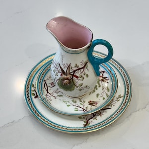 May include: A vintage porcelain creamer with a teal handle and a pink interior, resting on a matching saucer and plate. The set features a bird and branch design with gold trim and teal accents. The creamer and plates are white.