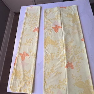 May include: Two yellow floral patterned fabric panels. The fabric features a repeating pattern of yellow and orange flowers and foliage. The panels are rectangular and appear to be folded. The fabric is likely used for curtains or other home decor.