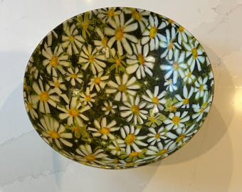 May include: A round bowl featuring a dense, repeating pattern of white daisy flowers with yellow centers set against a dark green, leafy background. The interior surface of the bowl is fully covered in this floral design, creating a continuous botanical motif across the entire concave surface of the vessel.