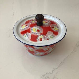 May include: A red and white enamelware bowl with a floral pattern and a dark wooden knob on the lid. The bowl has a white rim with a blue trim. A vintage kitchenware item.