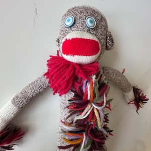 May include: A handmade sock monkey doll with a brown and white sock body, blue button eyes, and a red mouth. The monkey has a red yarn scarf and colorful yarn accents on its chest and arms. The arms and legs are made from socks.