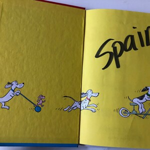 May include: Open book with illustrations of cartoon dogs on yellow pages. One dog pulls a small dog on a wheeled toy, another runs, and a third rides a scooter. The word "Spain" is written in large black letters.