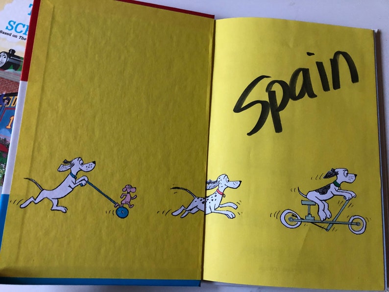 May include: Open book with illustrations of cartoon dogs on yellow pages. One dog pulls a small dog on a wheeled toy, another runs, and a third rides a scooter. The word "Spain" is written in large black letters.