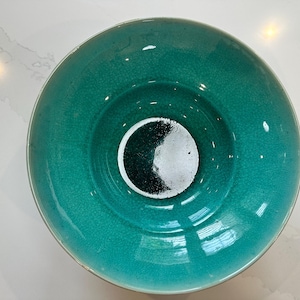 May include: A teal-colored ceramic bowl with a textured surface. The bowl has a circular design in the center, featuring a black and white pattern. The bowl's rim is slightly raised, and the overall shape is round.