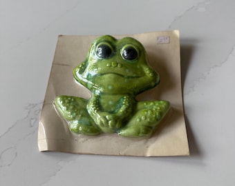 May include: A green ceramic frog ornament with large black eyes and a textured surface. The frog is attached to a piece of cardboard. The figurine has a glossy finish and is likely a decorative item.