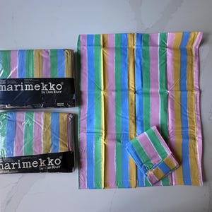 May include: A collection of Marimekko by Dan River bedding items. The set includes two wrapped packages, a large sheet, and a pillowcase, all featuring a colorful striped pattern in pink, yellow, blue, and green.