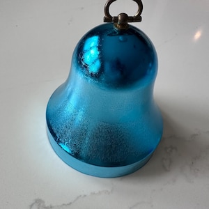 May include: A turquoise-blue bell-shaped ornament with a textured surface and a brass-coloured ring at the top. The bell has a rounded base and a smooth, reflective finish, suggesting a metallic or glass material.