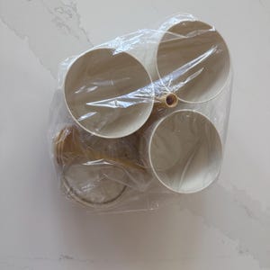 May include: A set of four cream-coloured ceramic bowls with a wooden handle, packaged in clear plastic. The bowls are round and appear to be stacked. The background is a white marble-patterned surface.