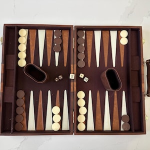 May include: Open backgammon board with brown and cream-coloured game pieces, dice, and leather dice cups. The board is open, revealing the playing surface with alternating light and dark triangles. The game is set up and ready to play.