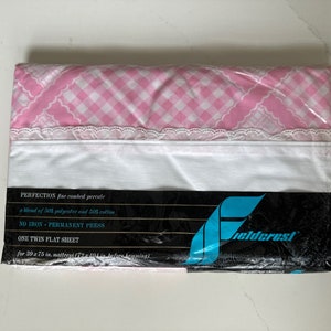 May include: A packaged twin flat sheet. The top portion features a pink and white gingham pattern with a white lace trim. The lower section is white. The packaging reads "PERFECTION fine combed percale" and "Fieldcrest".