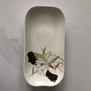 Peut inclure: Plat rectangulaire crème avec un motif floral. Le plat présente une fleur blanche avec des accents marron et verts. Le plat est en céramique et a des coins arrondis.
