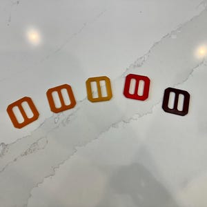 May include: Five colourful rectangular plastic buckles in shades of orange, yellow, red, and brown. Each buckle has two parallel slots. The buckles are arranged diagonally on a white marble surface.