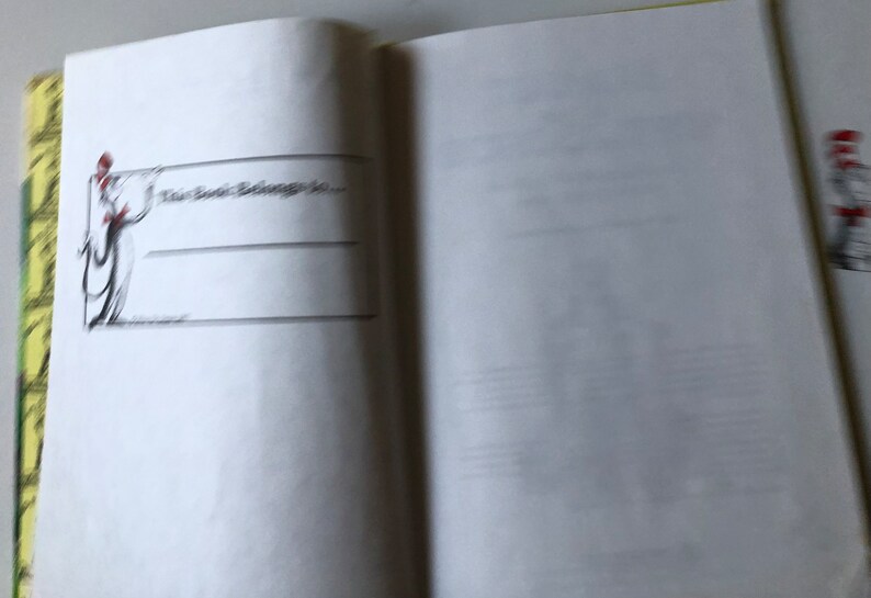 May include: Open book with a page featuring the text "This Book Belongs To..." and lines for writing. A black and white illustration of a figure holding a banner is on the left side of the page. The book's edge has a yellow and green pattern.