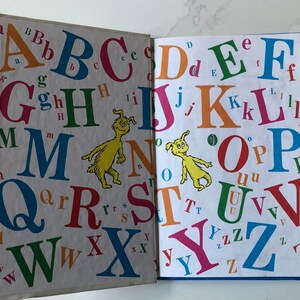 May include: Open book with colorful uppercase and lowercase letters of the alphabet. The letters are in various colors and sizes, with a yellow cartoon character on each page. The book is open to two pages.