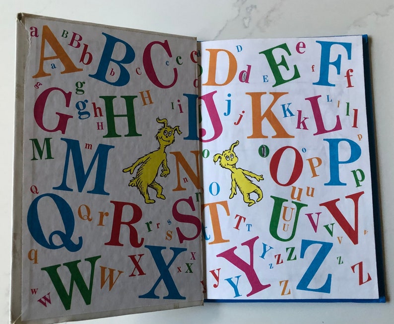 May include: Open book with colorful uppercase and lowercase letters of the alphabet. The letters are in various colors and sizes, with a yellow cartoon character on each page. The book is open to two pages.