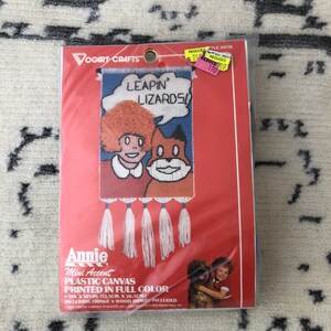 May include: A packaged Annie mini accent plastic canvas kit. The kit features a colorful design with the text "LEAPIN' LIZARDS" and illustrations of Annie and a dog. The package includes fringe and a wood dowel. The dimensions are 12.5cm by 26.5cm.