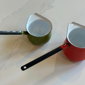 May include: Two small enamel saucepans with long black handles and white interiors. One saucepan is olive green and the other is bright red. Both feature a distinct pouring spout on the rim and a circular hole at the end of each handle.