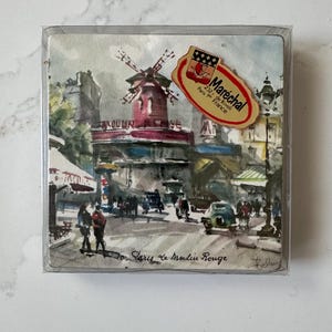 May include: A set of square coasters featuring a watercolor painting of the Moulin Rouge in Paris. The iconic red windmill is prominent, with figures and vehicles on the street below. The coasters are in a clear plastic case.