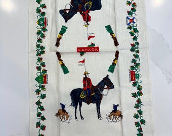 May include: White linen tea towel featuring a mounted Royal Canadian Mounted Police officer and horses. The towel has a border of green leaves and red berries, with Canadian flags and crests. The word "CANADA" is printed in red.